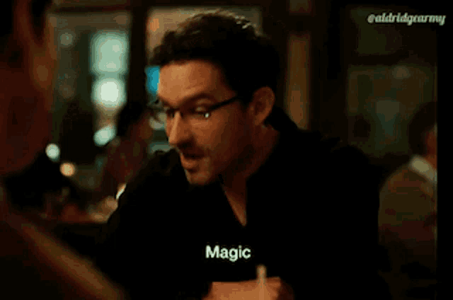 a man wearing glasses is sitting at a table talking to another man and saying `` magic '' .
