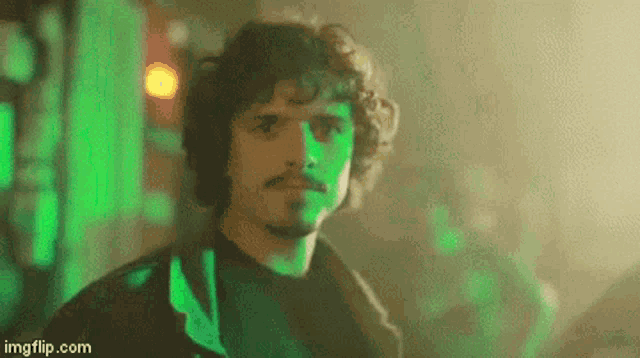 a man with curly hair and a mustache is standing in a dark room with a green light behind him .