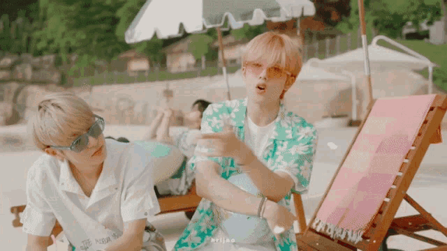 two young men are sitting on a beach under an umbrella and one is wearing sunglasses