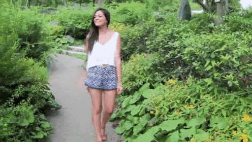 a woman wearing shorts and a white tank top is walking down a path in a garden .