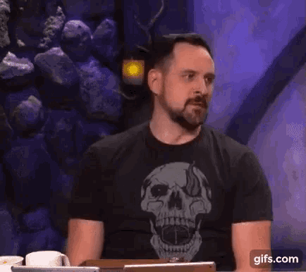 a man with a beard is wearing a black t-shirt with a skull on it and sitting at a table .