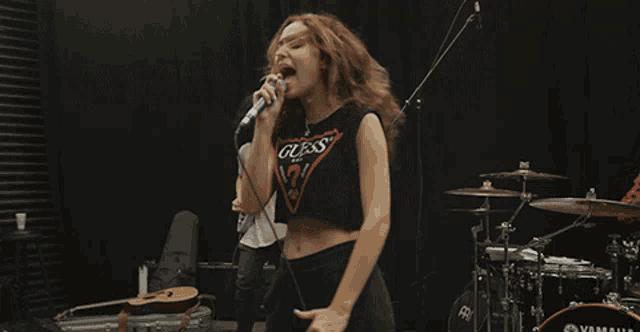 a woman wearing a guess shirt is singing in front of a drum set