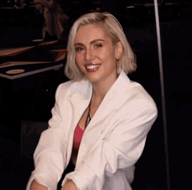a blonde woman wearing a white jacket and a pink top is smiling