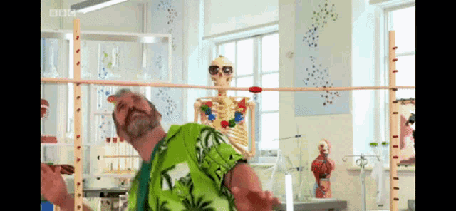 a man in a green shirt stands in front of a skeleton in a lab with bbc written on the screen