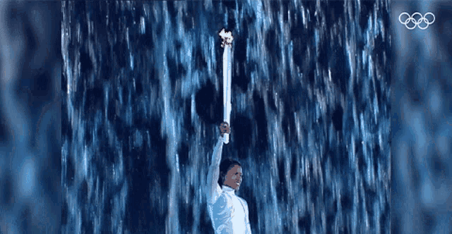 a person holding a torch in front of a waterfall with the olympic rings in the background
