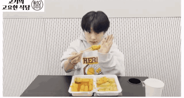 a young boy is sitting at a table eating food with chopsticks and wearing a hoodie that says erman