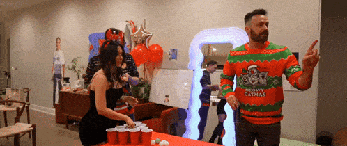 a man wearing a meow catmas sweater stands next to a woman playing beer pong