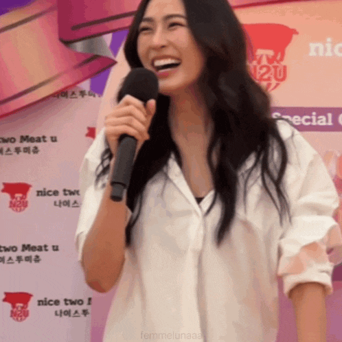 a woman laughs while holding a microphone in front of a sign that says " nice two "