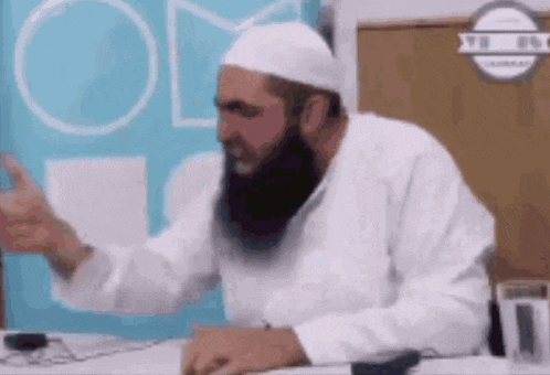 a man with a beard and a white hat is sitting at a desk with his hands outstretched .