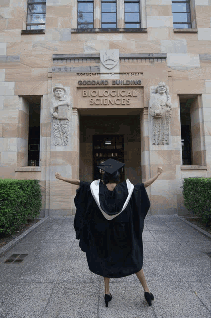 a woman in a graduation gown stands in front of the goddard building biological sciences