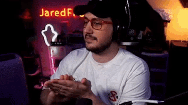 a man wearing headphones and glasses is clapping his hands in front of a neon sign that says jared f .