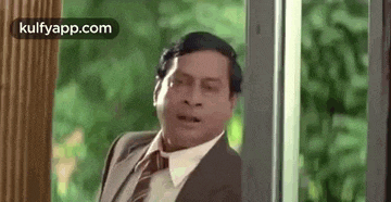a man in a suit and tie is standing next to a window and making a funny face .