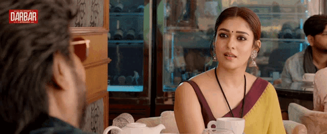 a woman in a sari sits at a table with a cup of coffee in front of a sign that says ' darbar '