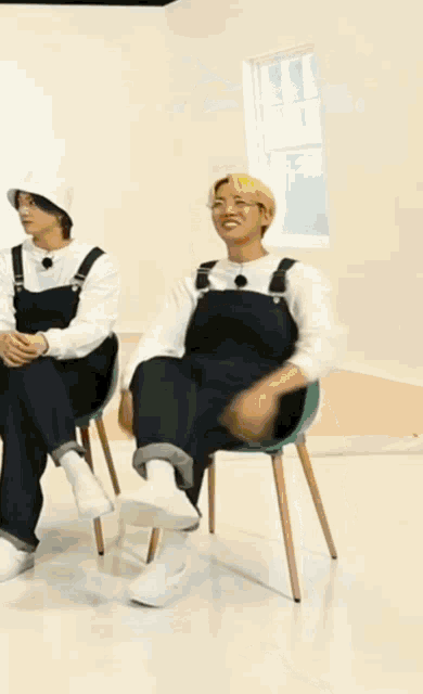 a man in overalls is laying on a chair while another man sits next to him