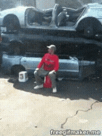 a man in a red shirt is sitting on a bucket in front of a pile of cars ..