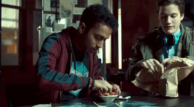 two men are sitting at a table eating food from a paper bag