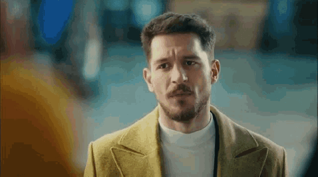a man with a beard wearing a yellow coat and a white turtleneck is looking at the camera .