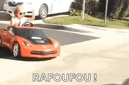 a young boy is driving a red toy car on a street .