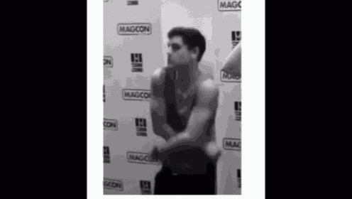 a man without a shirt is standing in front of a wall with magcon on it .