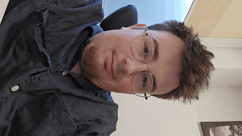 a man wearing glasses and a denim shirt looks down at the camera