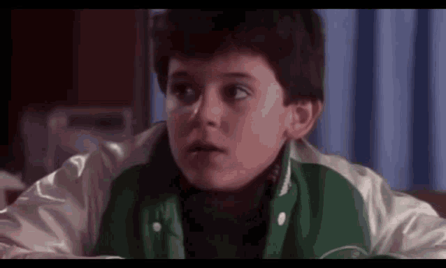 a young boy wearing a green and white jacket is sitting on a bed .