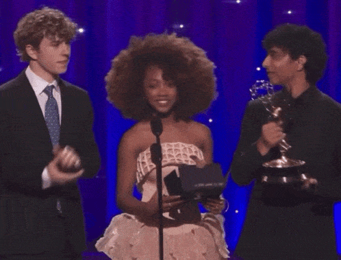 a woman stands in front of a microphone with two men behind her
