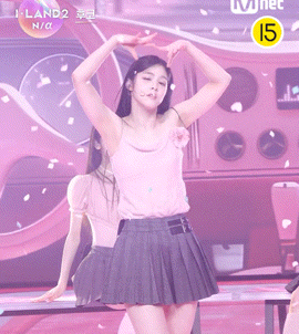 a girl in a pink tank top and gray pleated skirt is dancing on a stage in front of a mnet logo