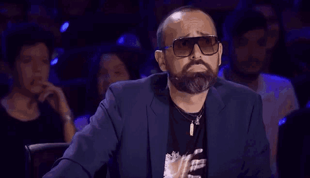 a man with a beard wearing sunglasses and a suit is sitting in a theatre .