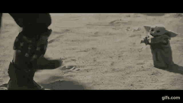 a baby yoda is standing in the sand next to a person in a desert .