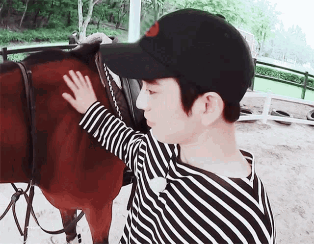 a man wearing a black hat and striped shirt petting a brown horse