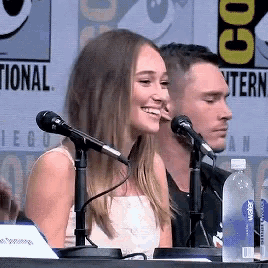a woman sits at a table with two microphones and a bottle of aquafina water in front of her