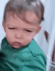 a baby in a green shirt is making a funny face and looking at the camera .
