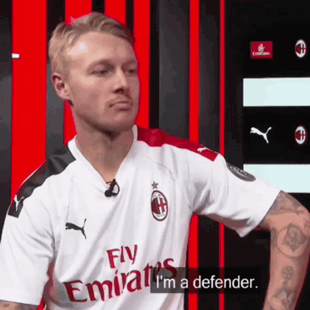 a man wearing a fly emirates shirt is standing in front of a red and black wall