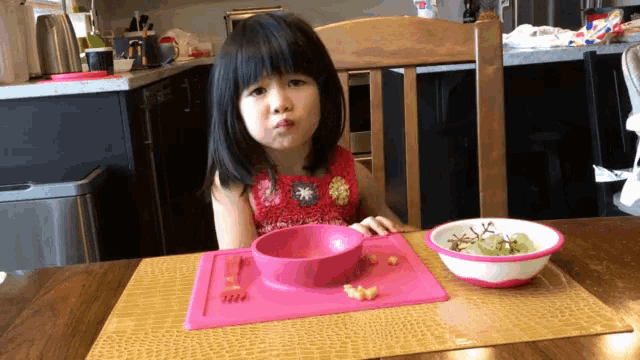 a little girl is sitting at a table with a bowl of food and a plate of food