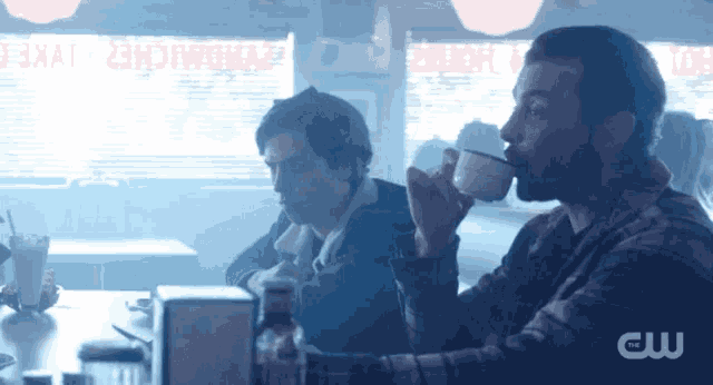 two men are sitting at a table in a diner drinking from cups .