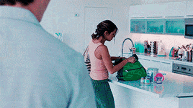 a woman in a pink tank top is standing in a kitchen holding a green bag