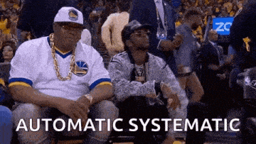 two men are sitting in the stands at a basketball game . one of the men is wearing a golden state warriors jersey .