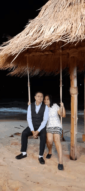 a man and woman sit on a swing under a thatched roof on the beach