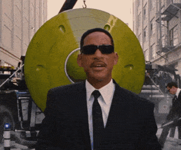 a man in a suit and tie wearing sunglasses stands in front of a green object
