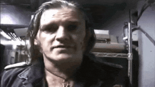 a man with long hair and a necklace is standing in a dark room and looking at the camera .