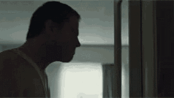a man in a tank top is standing in a dark room looking out of a window .
