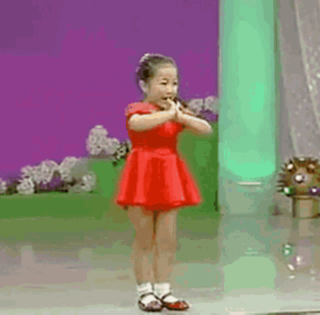 a little girl in a red dress is clapping her hands in front of a green pillar with chinese writing on it .