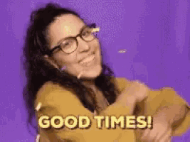 a woman wearing glasses and a yellow sweater is smiling and says `` good times '' .