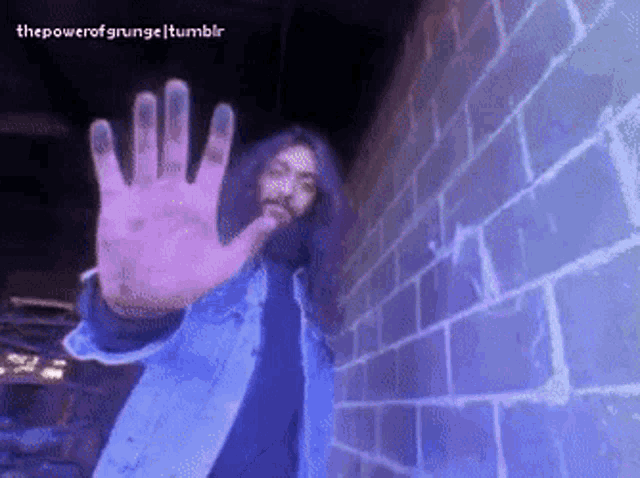 a man with long hair and a beard is standing in front of a brick wall with his hand outstretched ..