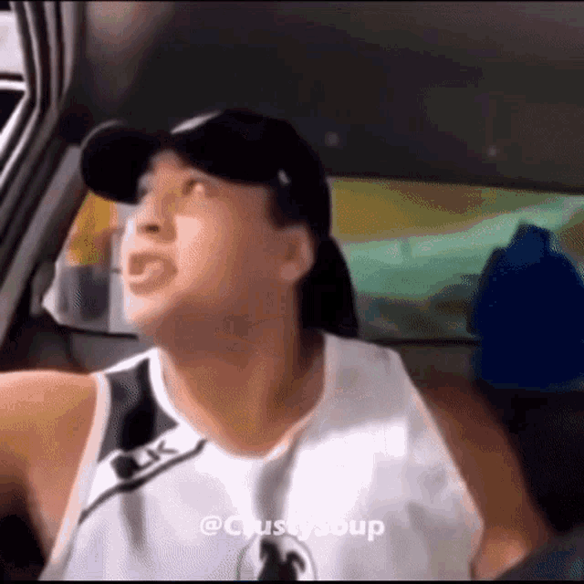 a man wearing a black hat and a white tank top is driving a car