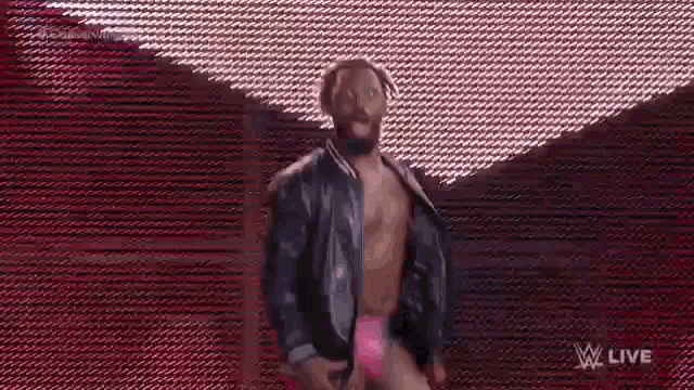 a man in a leather jacket and pink shorts is standing on a stage in front of a red wall .