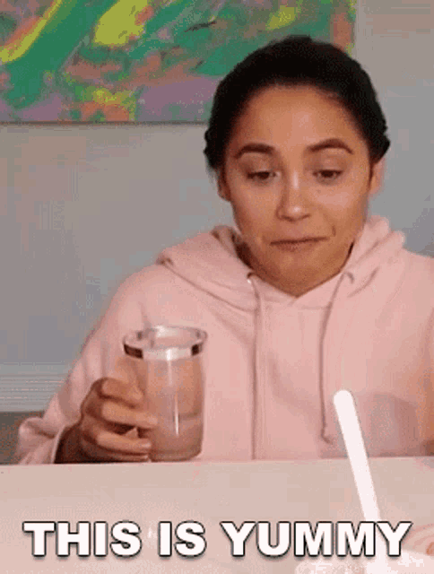a woman in a pink hoodie is sitting at a table holding a glass of water and a spoon .