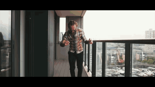 a man in a plaid shirt stands on a balcony overlooking the city
