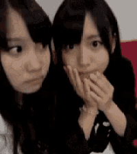 two girls are sitting next to each other on a red couch and covering their mouths with their hands .