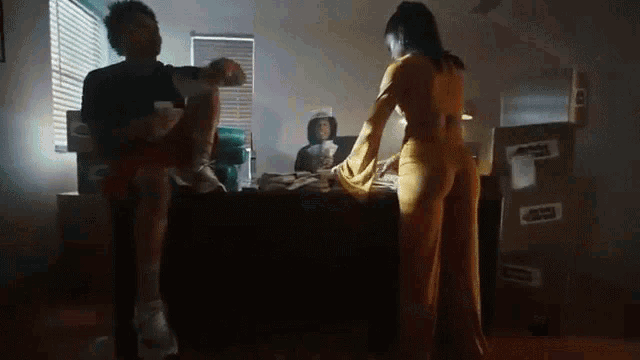 a woman in a yellow dress is standing next to a man sitting on a bed in a room with boxes .